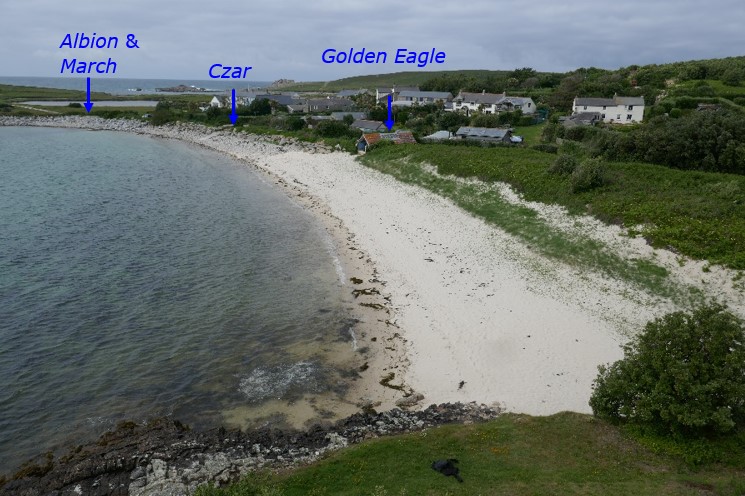 A sandy beach curving along a bay with calm, clear water, backed by green fields and scattered buildings. Blue text labels mark locations associated with historic pilot gig sheds, including Albion & March, Czar, and Golden Eagle.