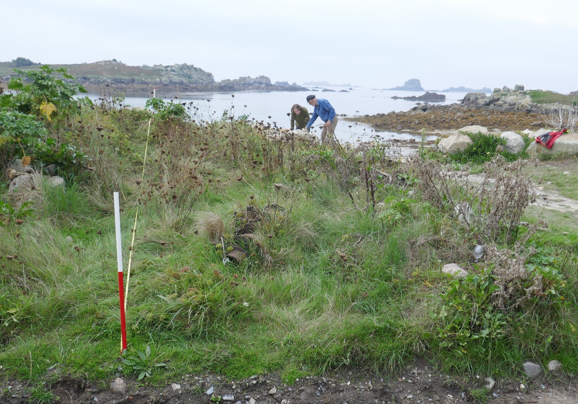 Archaeologists working near the shoreline, measuring and recording an archaeological site with a red-and-white survey pole and tape in the foreground. The landscape consists of grassy terrain, rocky outcrops, and calm tidal waters with small islands in the distance.