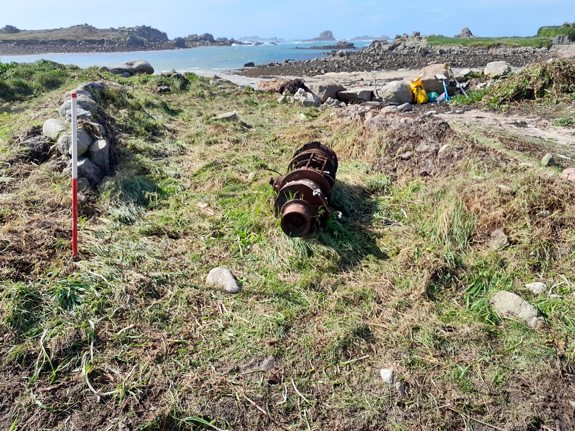  A red-and-white survey pole stands in a cleared area of grass and vegetation, with a rusted mechanical object lying nearby. The site overlooks a rocky shoreline.