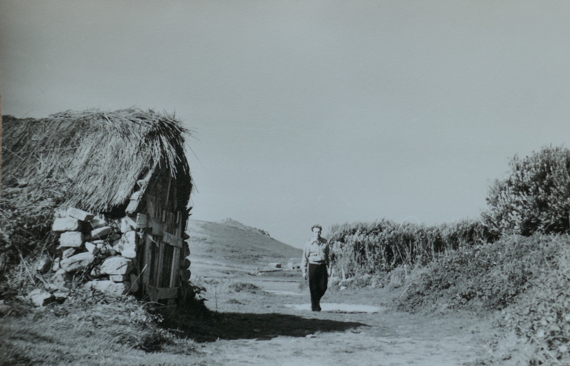 A 1960s black-and-white photograph of the thatched Czar gig shed with a wooden door, situated along a track. A person walks towards the camera, with a landscape of hedgerows and low hills in the background.