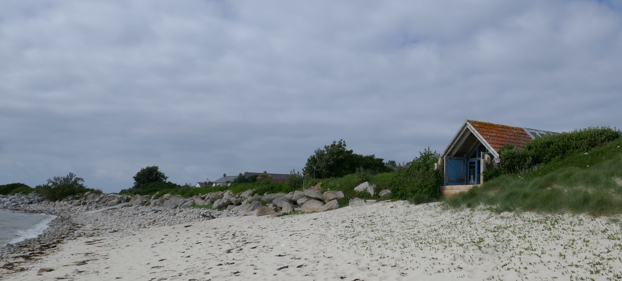 A restored gig shed, now a studio, with a pitched roof, blue doors, and weathered wooden beams, partially hidden by dunes and greenery. Large stones and boulders form sea defences along the edge of the sandy beach.