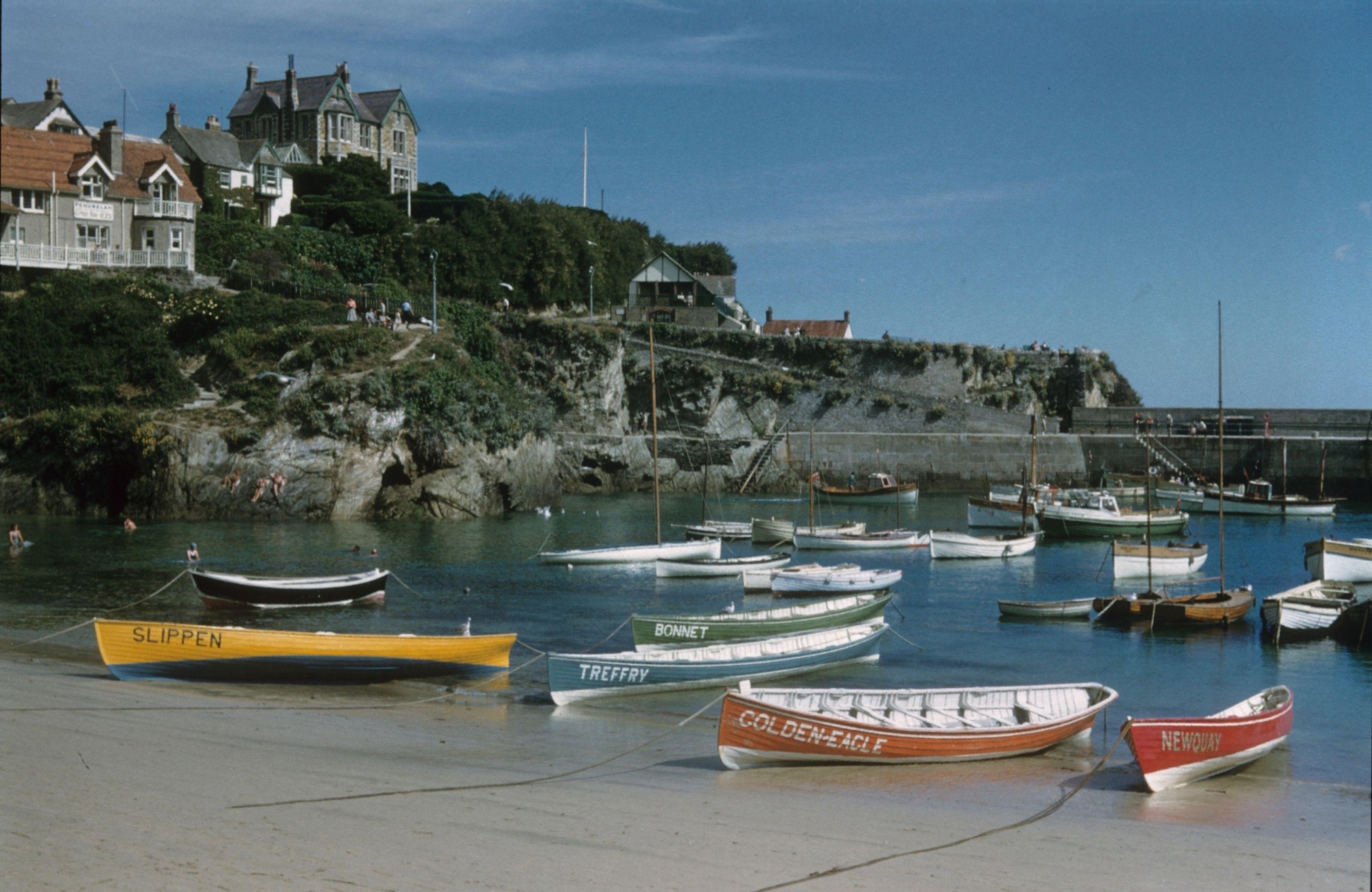 A 1956 photograph of Newquay Harbour showing several wooden pilot gigs on the sandy shore and in the water. In the foreground, the yellow gig Slippen, the green gig Bonnet, the white and blue gig Trefrry, the orange and white gig Golden Eagle, and the red gig Newquay are lined up. More small boats and sailing vessels float in the harbour, with a stone harbour wall, rocky cliffs, and houses overlooking the scene under a bright blue sky.