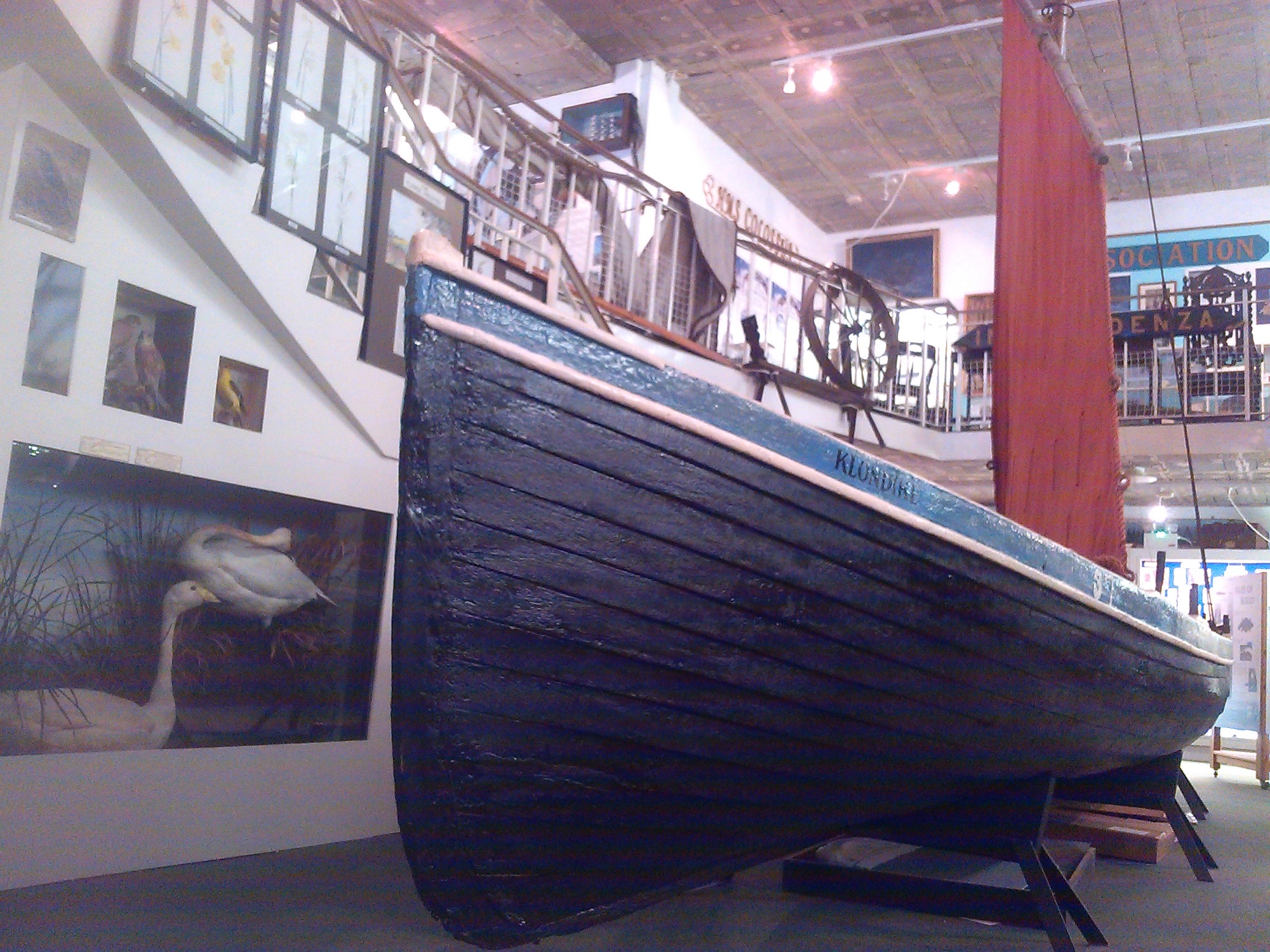 A historic wooden boat named Klondyke is displayed inside a museum, its dark blue hull resting on supports. It has a red sail. The background features maritime exhibits, framed pictures, a ship's wheel