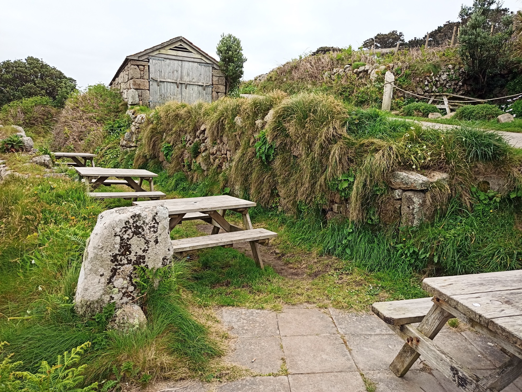 Sites of sheds, now grassy roofless walled platforms, once used for the Klondyke, now occupied by picnic tables. The granite shed now housing the gig Shah stands above; a slipway slants down left of it.