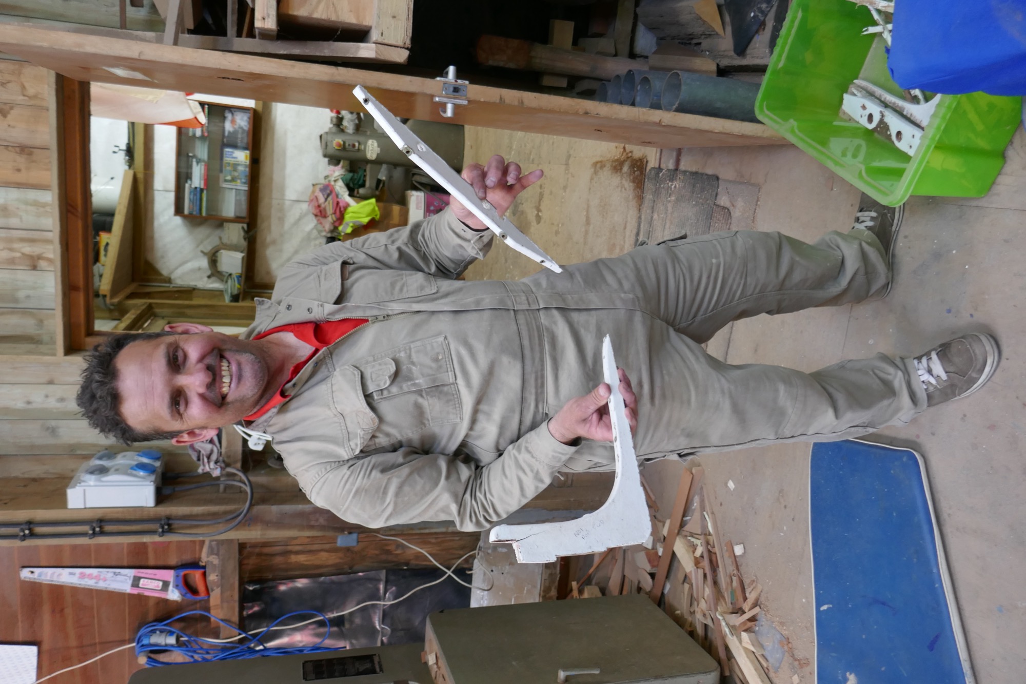 Boatbuilder Andrew Hicks stands in a workshop, holding two white-painted wooden gig components. The background features wooden walls, shelves with tools and supplies, and a workbench covered with materials, while wood shavings and offcuts are scattered on the floor.