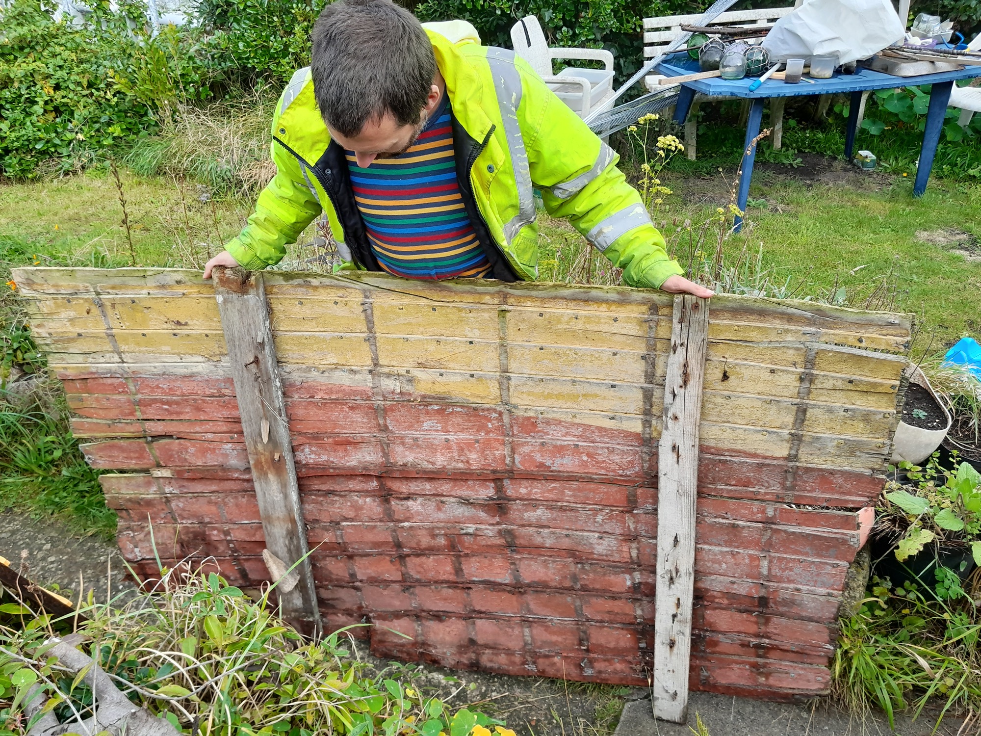 Harry Legg wearing a high-visibility jacket and a colourful striped jumper holds up a weathered wooden panel with faded yellow and red paint. The background features a grassy garden area with a blue outdoor table, white plastic chairs, and various gardening items scattered around.