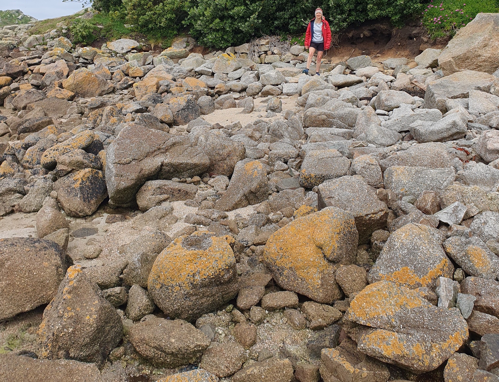 A rocky shoreline with large, lichen-covered boulders scattered across the terrain. Project lead Cathy wearing a red jacket stands at the edge of the rocks, looking down towards the uneven ground.