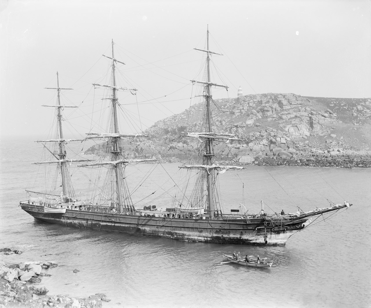 A historic black-and-white photograph of the three-masted sailing ship Horsa, grounded in shallow waters near a rocky coastline. In the foreground, a wooden rowing gig approaches the ship.