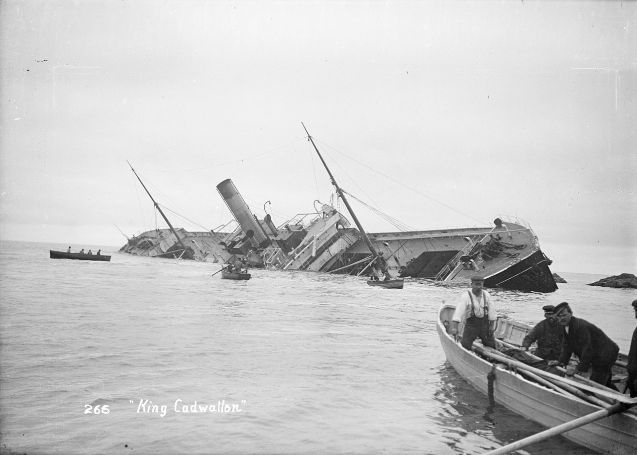 A black-and-white photograph of the wreck of the steamship King Cadwallon, which is partially submerged and listing heavily to one side near a rocky shoreline. Several gigs are in the foreground, with some men observing from a wooden gig while others row towards the wreckage.