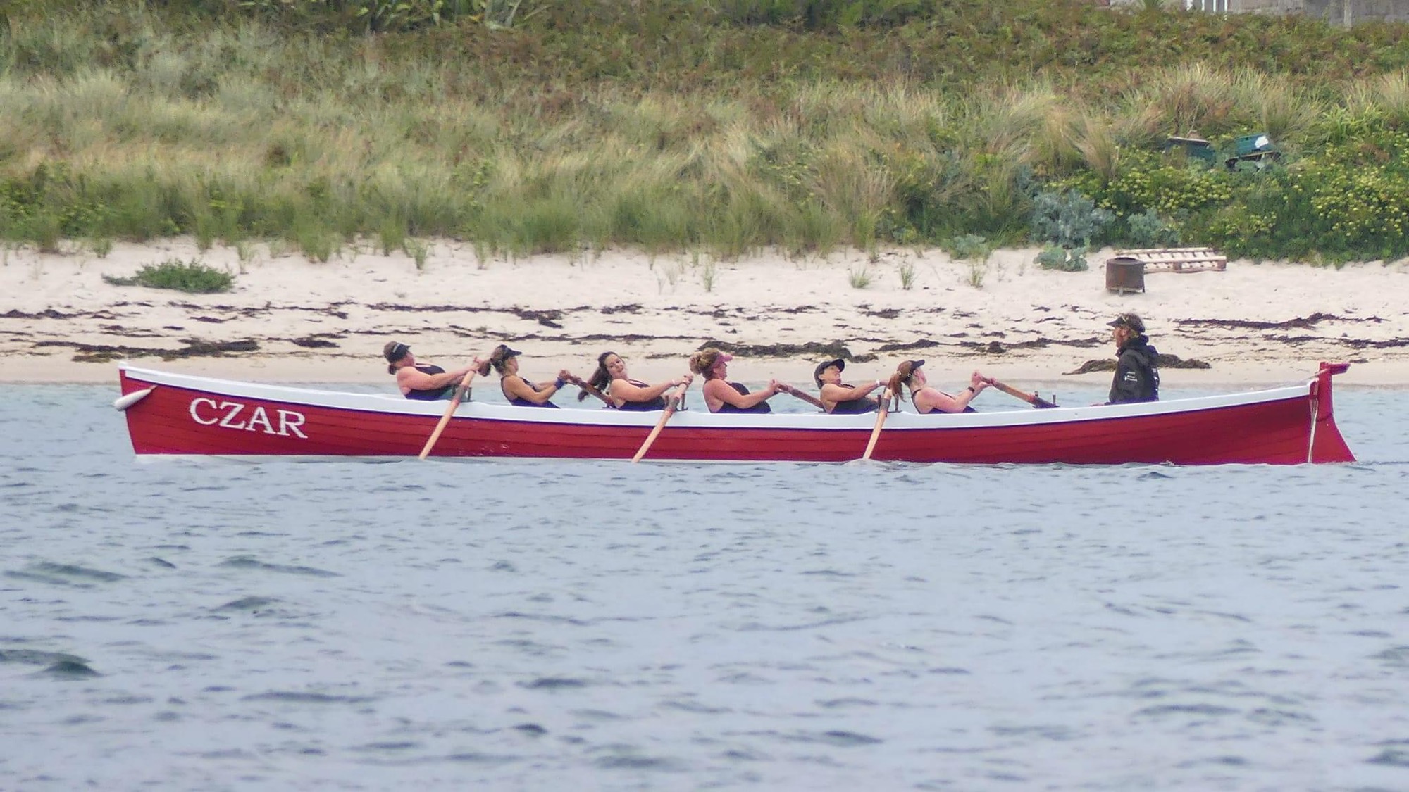 A red and white pilot gig named "Czar" is rowing through the water with a crew of six women pulling in unison. A coxswain stands at the stern, guiding the team as they row past a sandy beach with grass-covered dunes in the background.