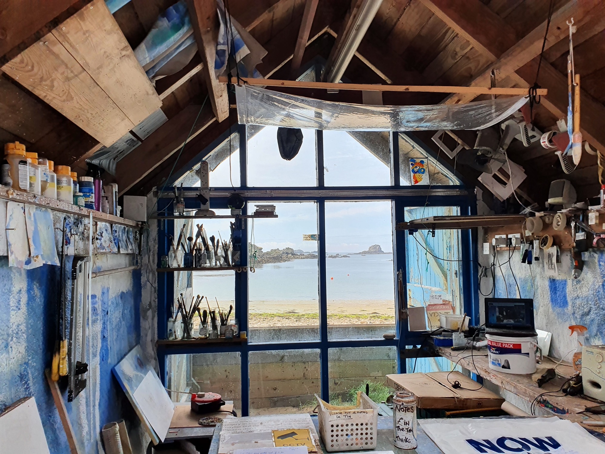 The interior of an artists studio with wooden beams and shelves filled with tools, brushes, and various supplies. A large window and open doorway frame a stunning seaside view with a sandy shore, calm waters, and small islands and rocks in the distance.