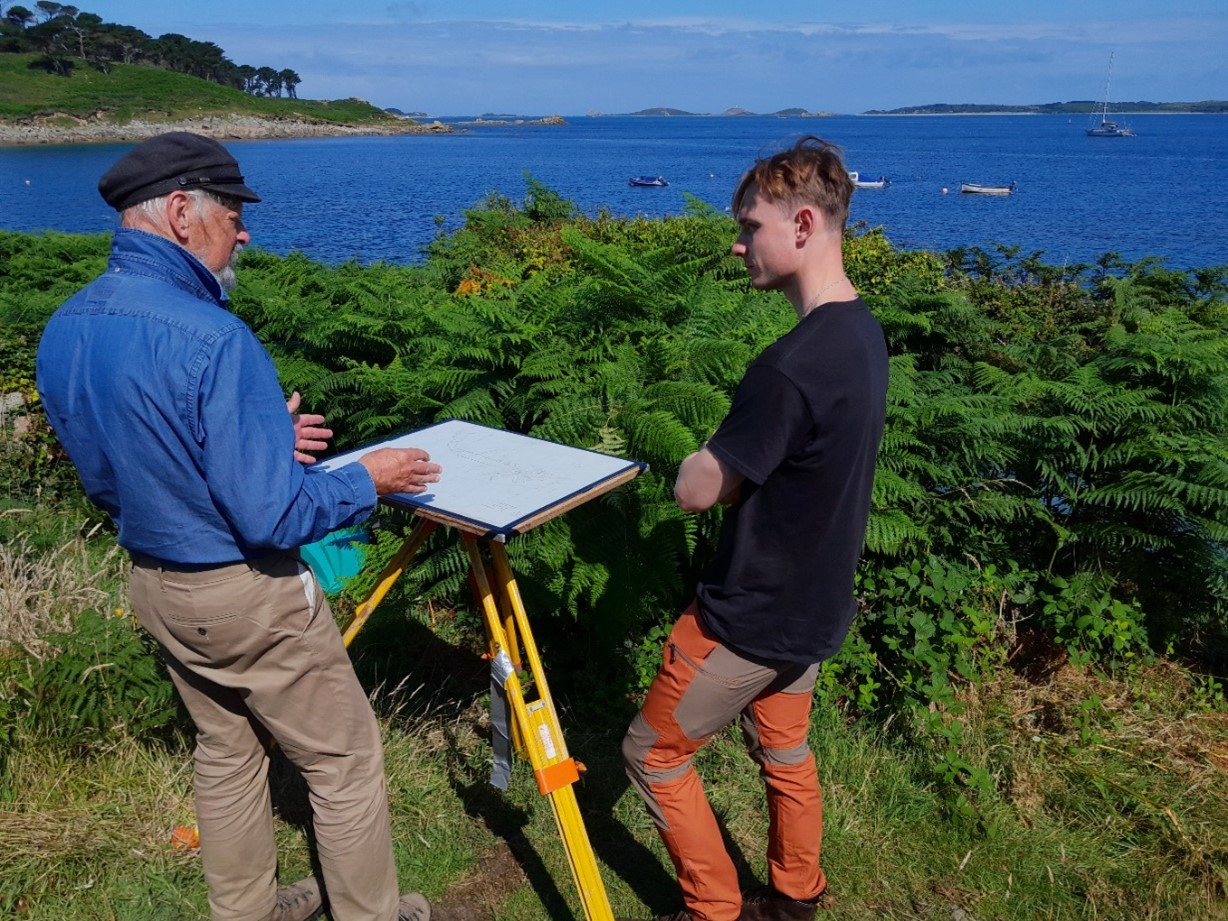 Mark and Ben stand beside a tripod-mounted drawing board, engaged in discussion during an outdoor survey. Behind them, lush green ferns frame a coastal scene with blue waters, moored boats, and distant islands under a clear sky.