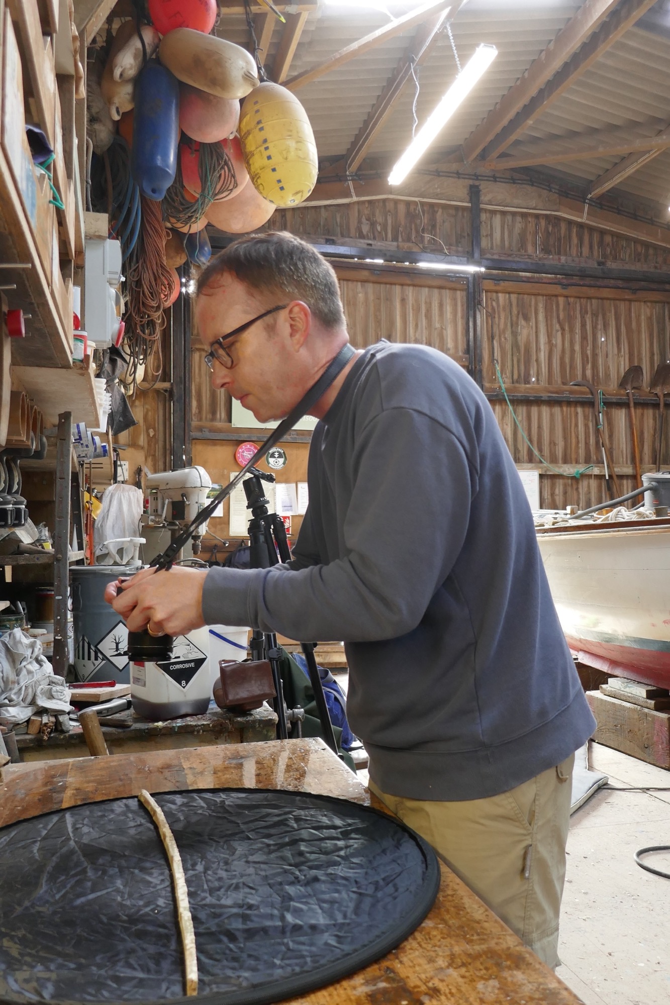 Tom photographs a thin wooden gig rib on a workbench inside a workshop. The background features wooden walls, hanging buoys, tools, and a partially visible boat under repair.
