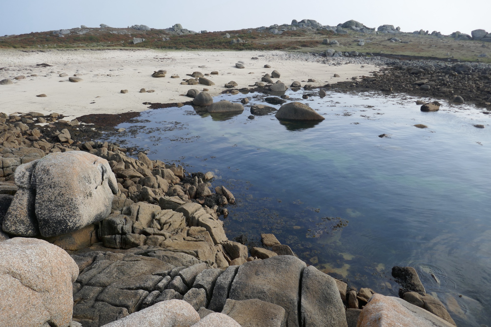 A coastal scene with a sandy beach and scattered granite boulders leading into clear, shallow water. In the foreground, weathered rock formations extend into the sea, while the background features a low hill dotted with more large granite outcrops.