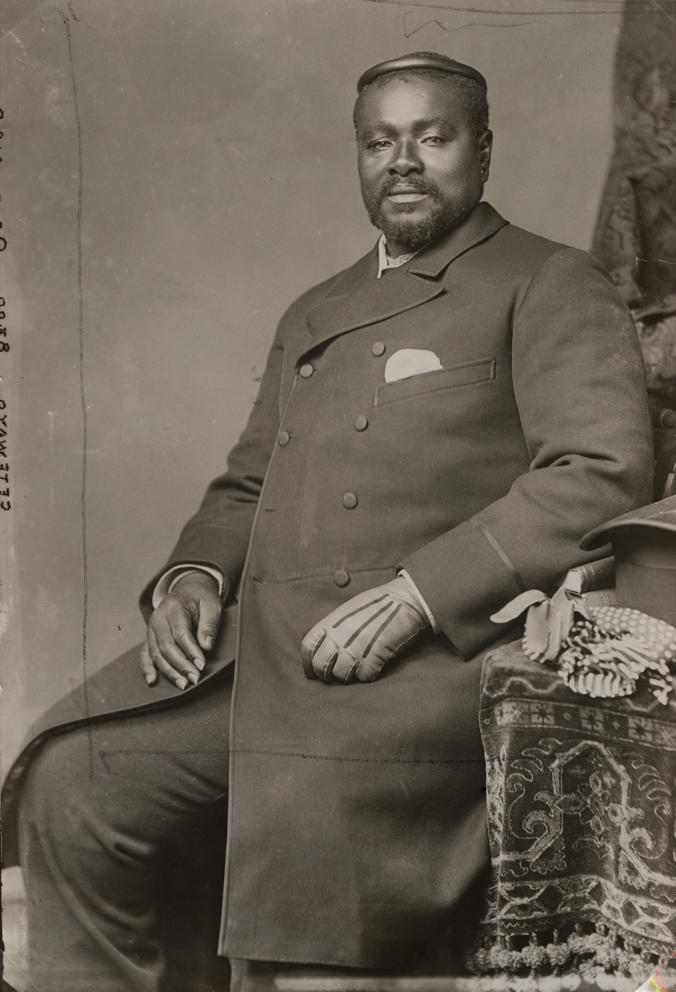 A historical black-and-white portrait of King Cetewayo, the Zulu leader, seated in formal attire. He wears a dark double-breasted coat with a pocket square and gloves, and on his head, he has a traditional Zulu isicoco, a woven or fitted headband-like headdress symbolising his status as a leader.