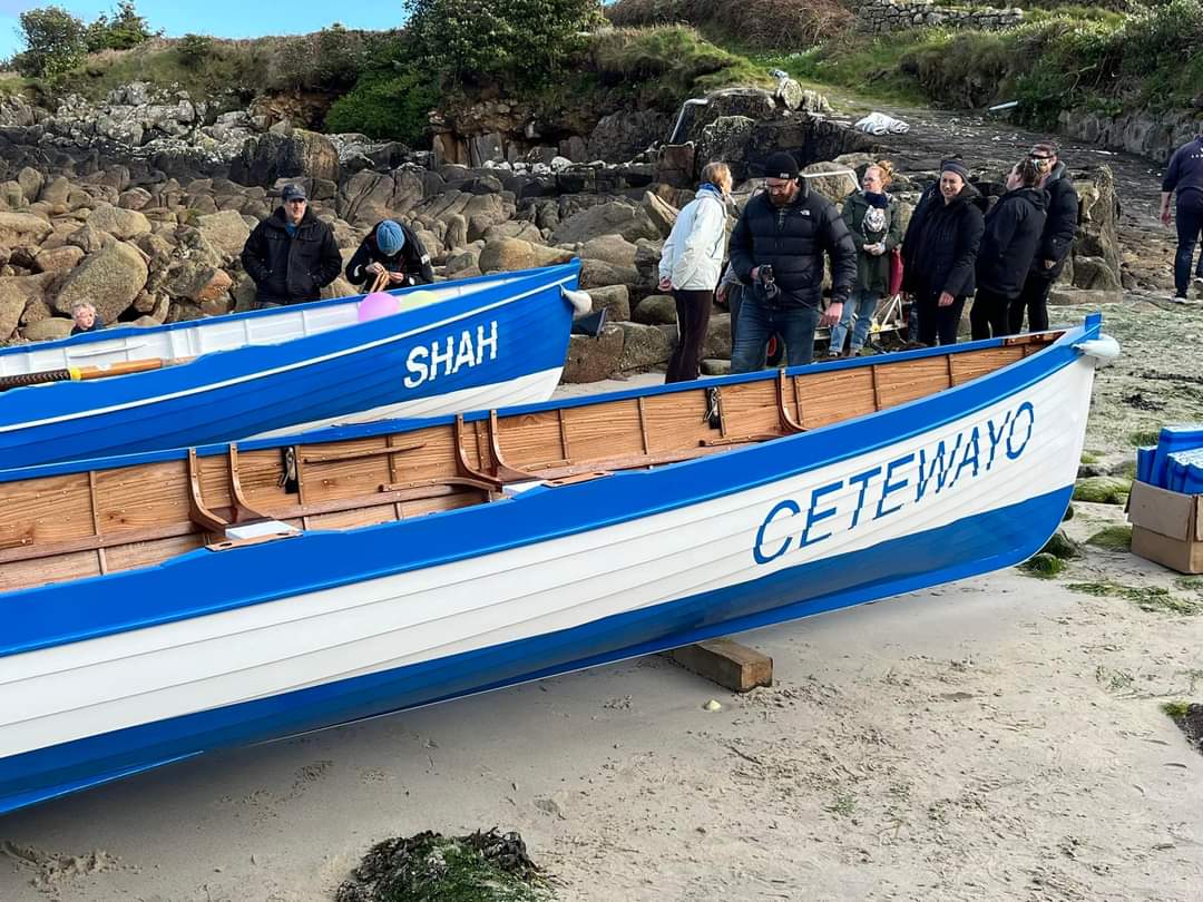 Two traditional wooden pilot gigs with blue and white paint, named Shah and Cetewayo, rest on the sand during a launch event. A group of people, dressed in warm clothing, stand nearby, with a rocky shoreline and grassy hillside in the background.