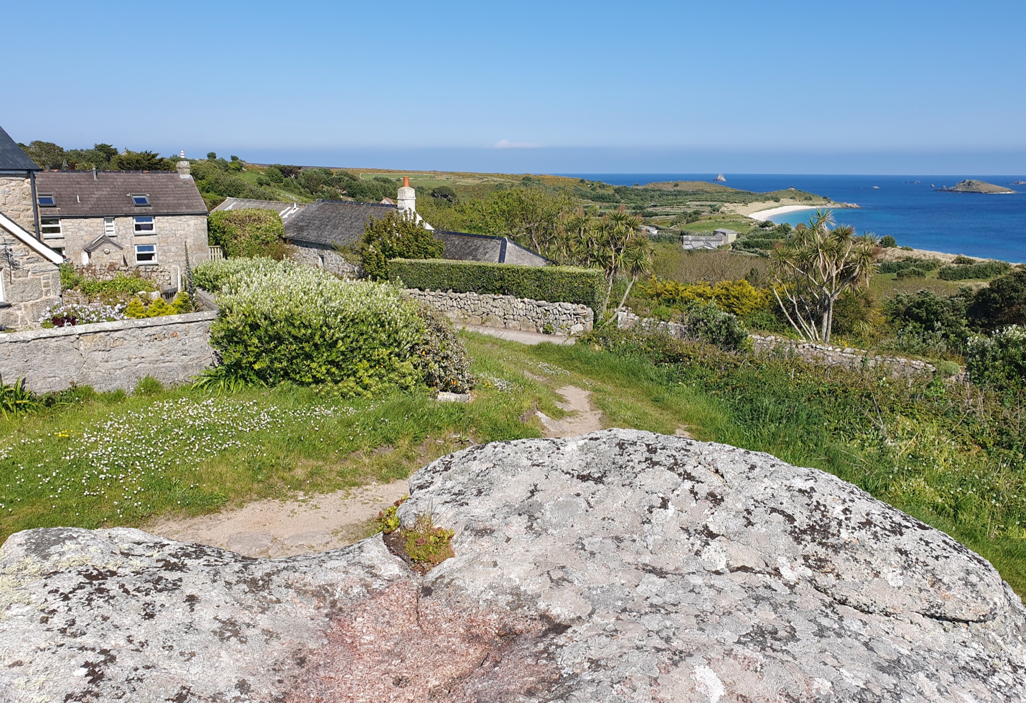 View from the top of Signal Rock looking down over stone cottages, hedgerows, and a winding path. In the background are palm trees, a sandy beach, blue sea, and small islands stretch out under a bright sky.