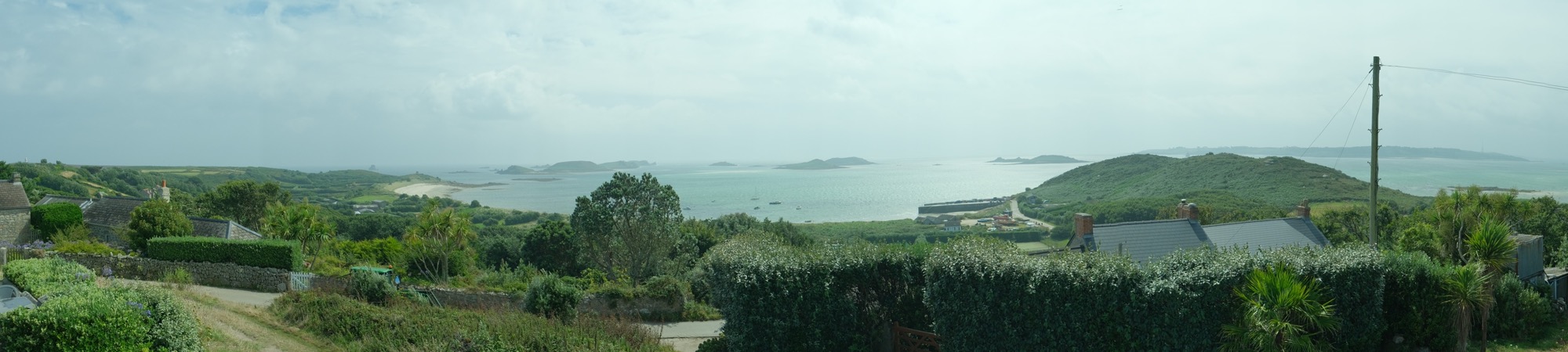 A panoramic view of St Martin’s, Isles of Scilly, showing rolling green fields, houses, and an expansive sea dotted with islands. The scene is framed by stone walls and hedgerows in the foreground.