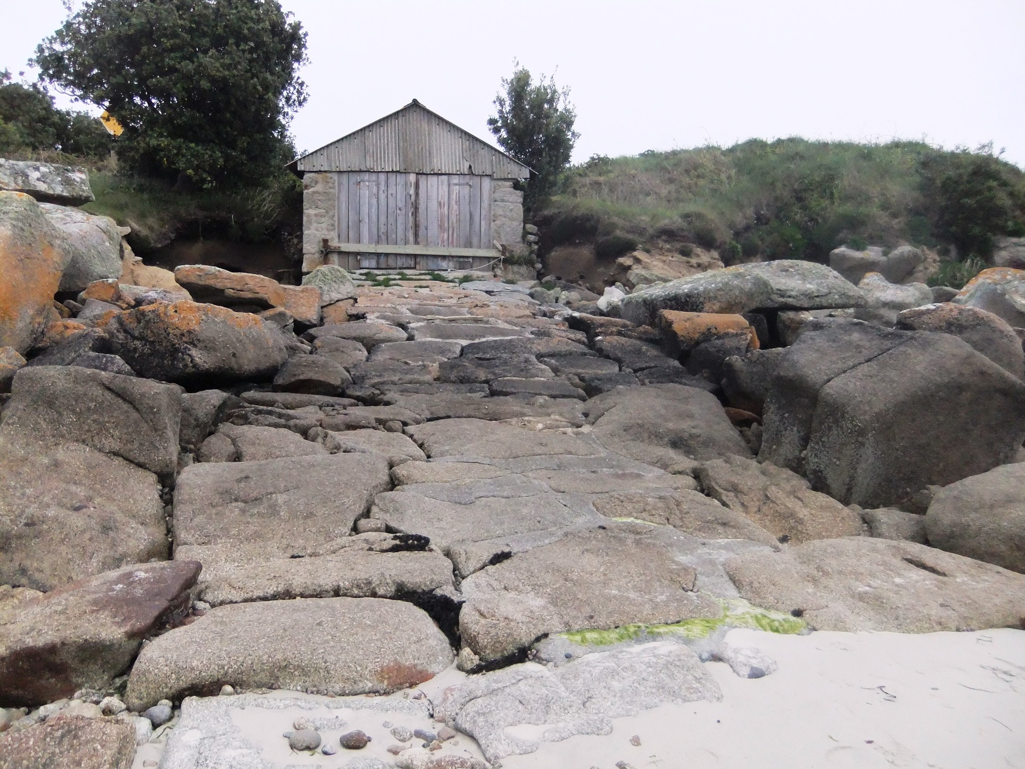 A historic stone slipway built from large, irregular granite blocks leading up to a weathered gig shed with wooden doors and a corrugated metal gable end. Moss and lichen cover the rocks, and the sandy beach is visible at the bottom.