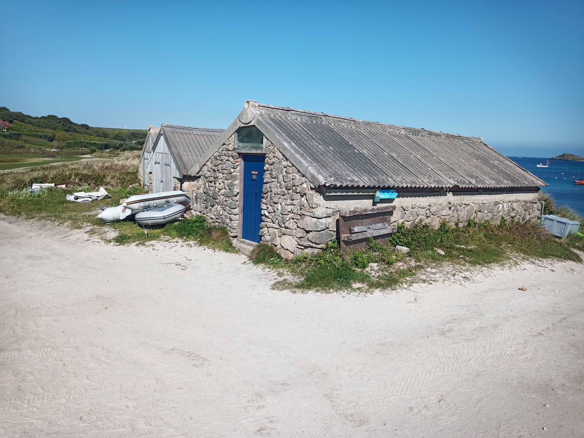 A row of traditional stone gig sheds with grey corrugated roofs, located near a sandy path by the sea. A blue wooden door stands out among the stonework, with small boats and equipment scattered outside.