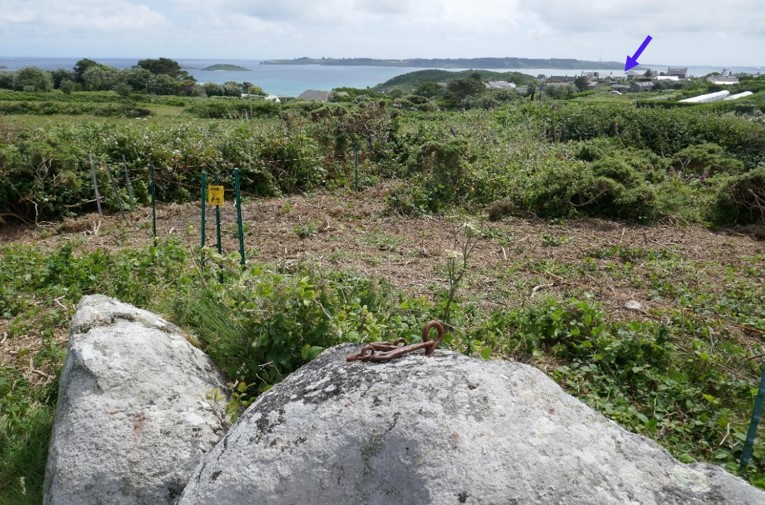 A granite rock with a rusted iron ring embedded on top, surrounded by low shrubs and vegetation. The view extends over green fields and the blue sea, with distant islands visible on the horizon.