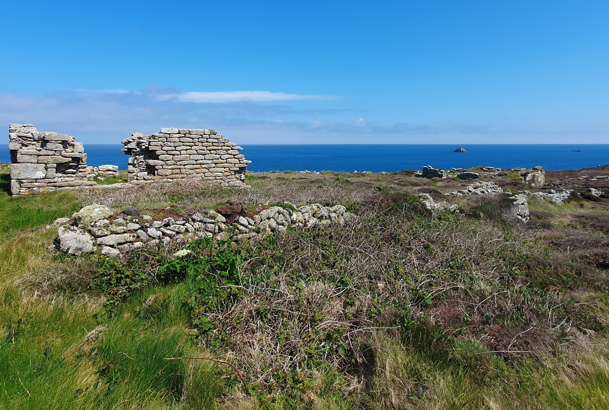 The ruins of an old stone structure on Chapel Down, overlooking the deep blue sea. The remains of the granite walls stand amid grassy, windswept terrain, with a rocky islet visible in the background.