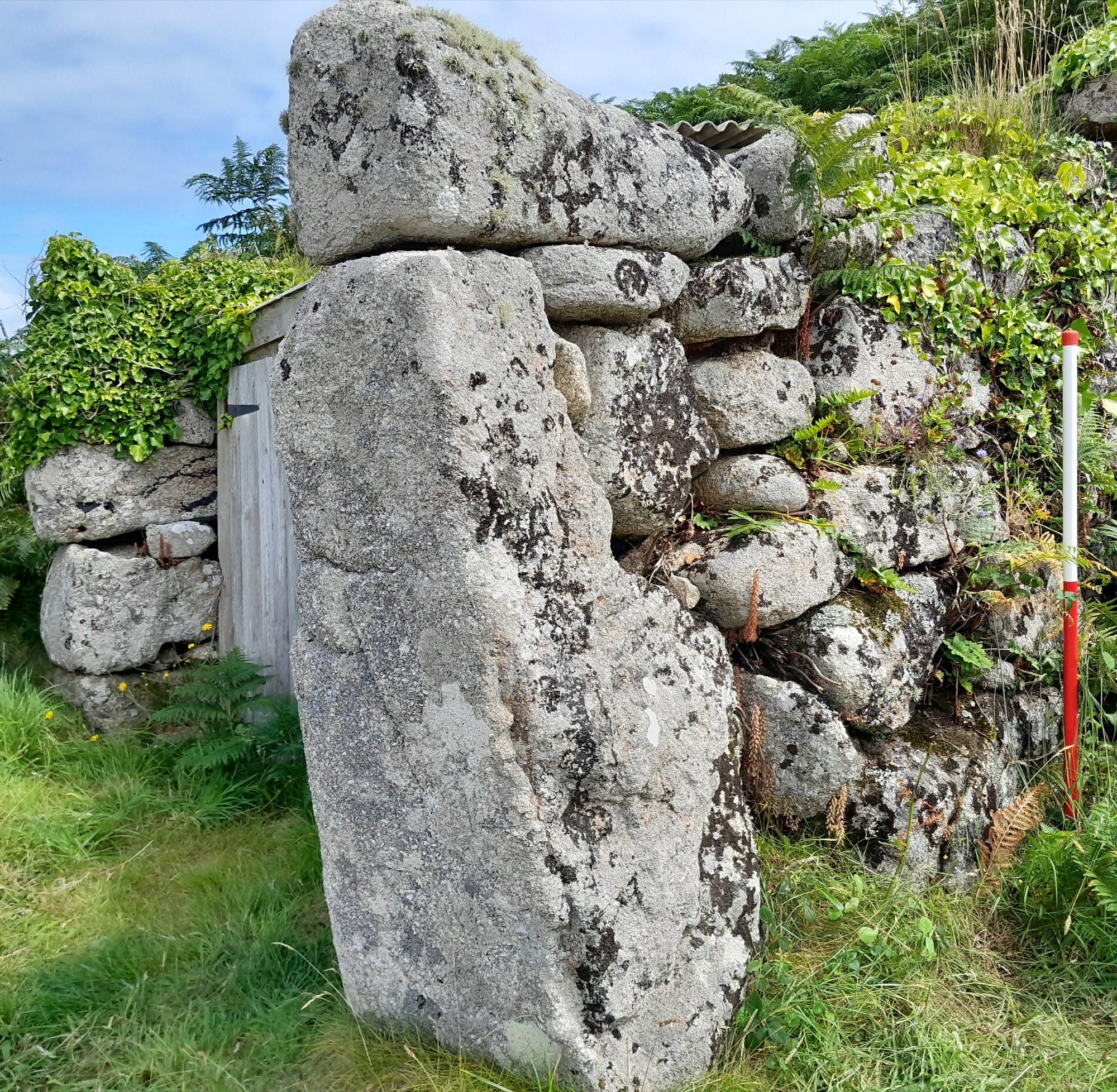 A dry-stone wall built from large, irregular granite blocks, with a tall vertical stone forming the side of a doorway. Vegetation is growing around the structure.