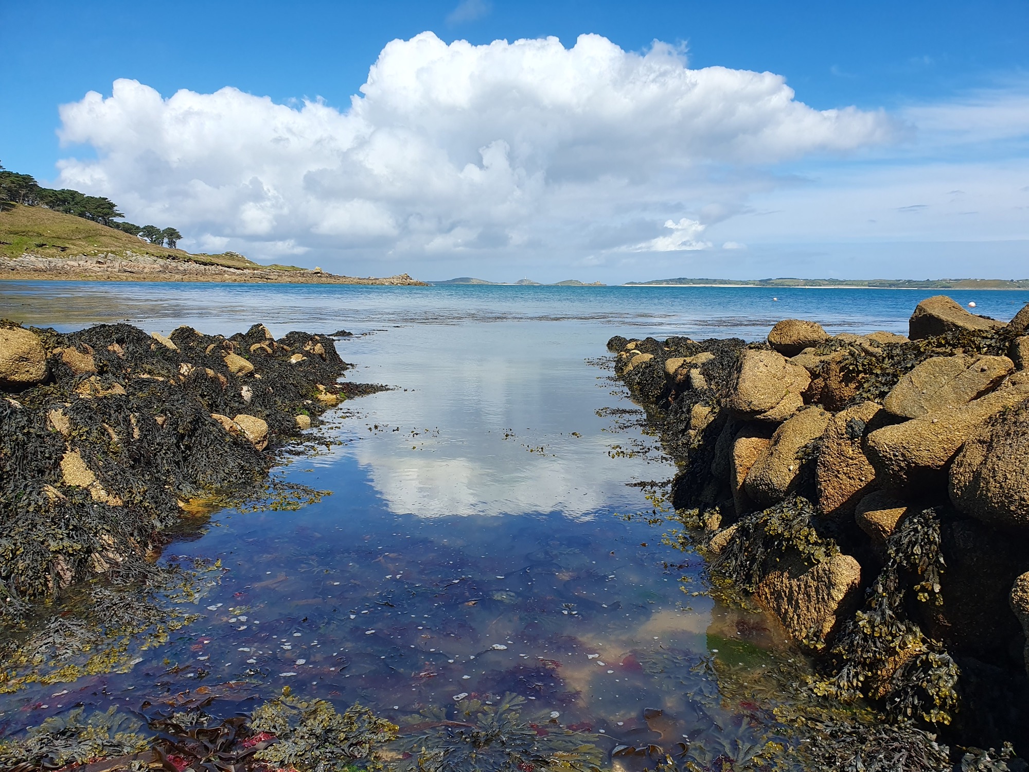 A channel leading to the sea, flanked by stone revetments covered in seaweed. The calm water reflects the blue sky and white clouds, with distant islands visible on the horizon.