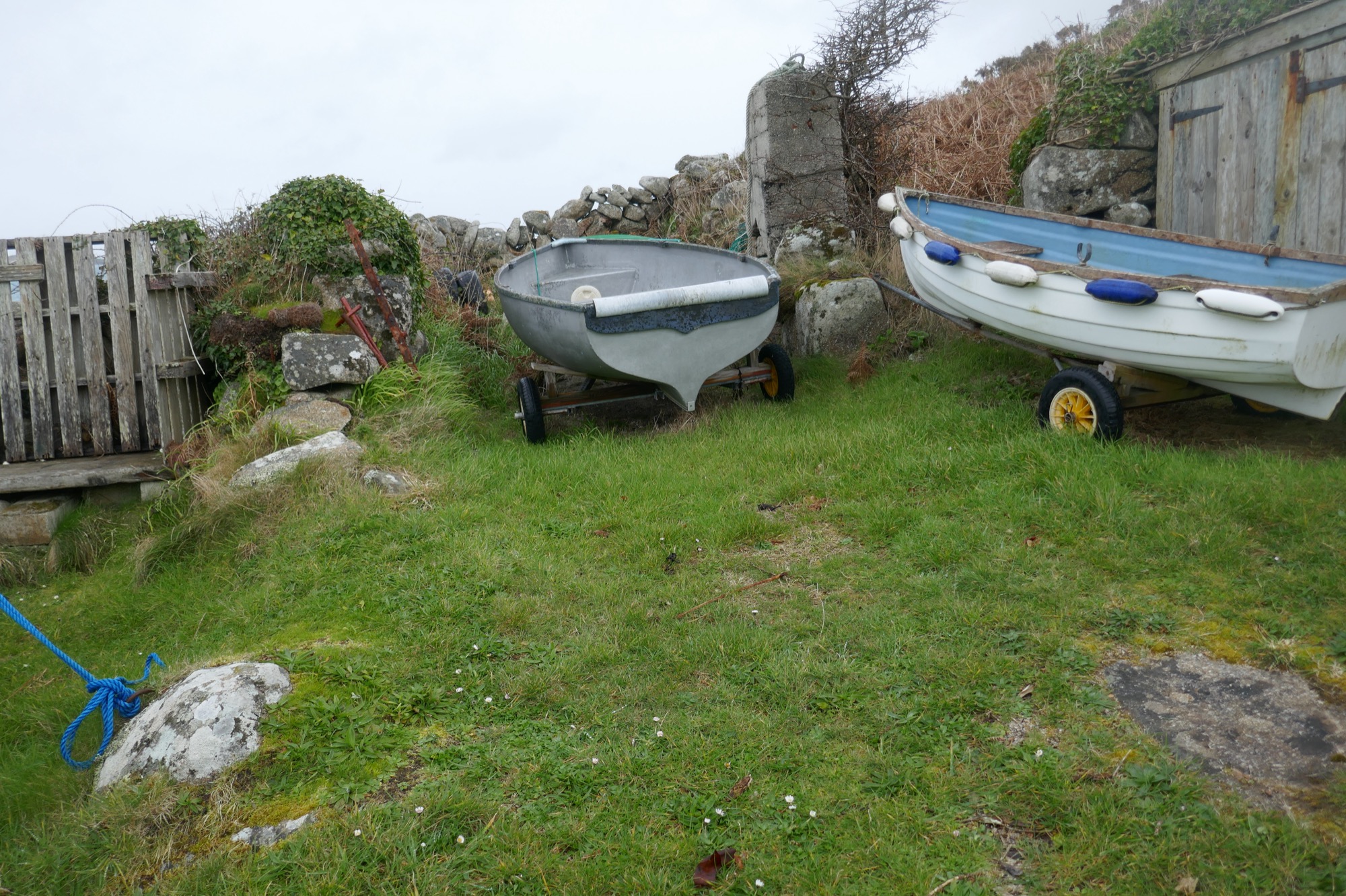 Two small boats on wheeled trailers rest on a grassy area near a wooden shed and a dry-stone wall. The left boat is grey and fibreglass, while the right boat is white with blue trim and has fenders attached to its side.