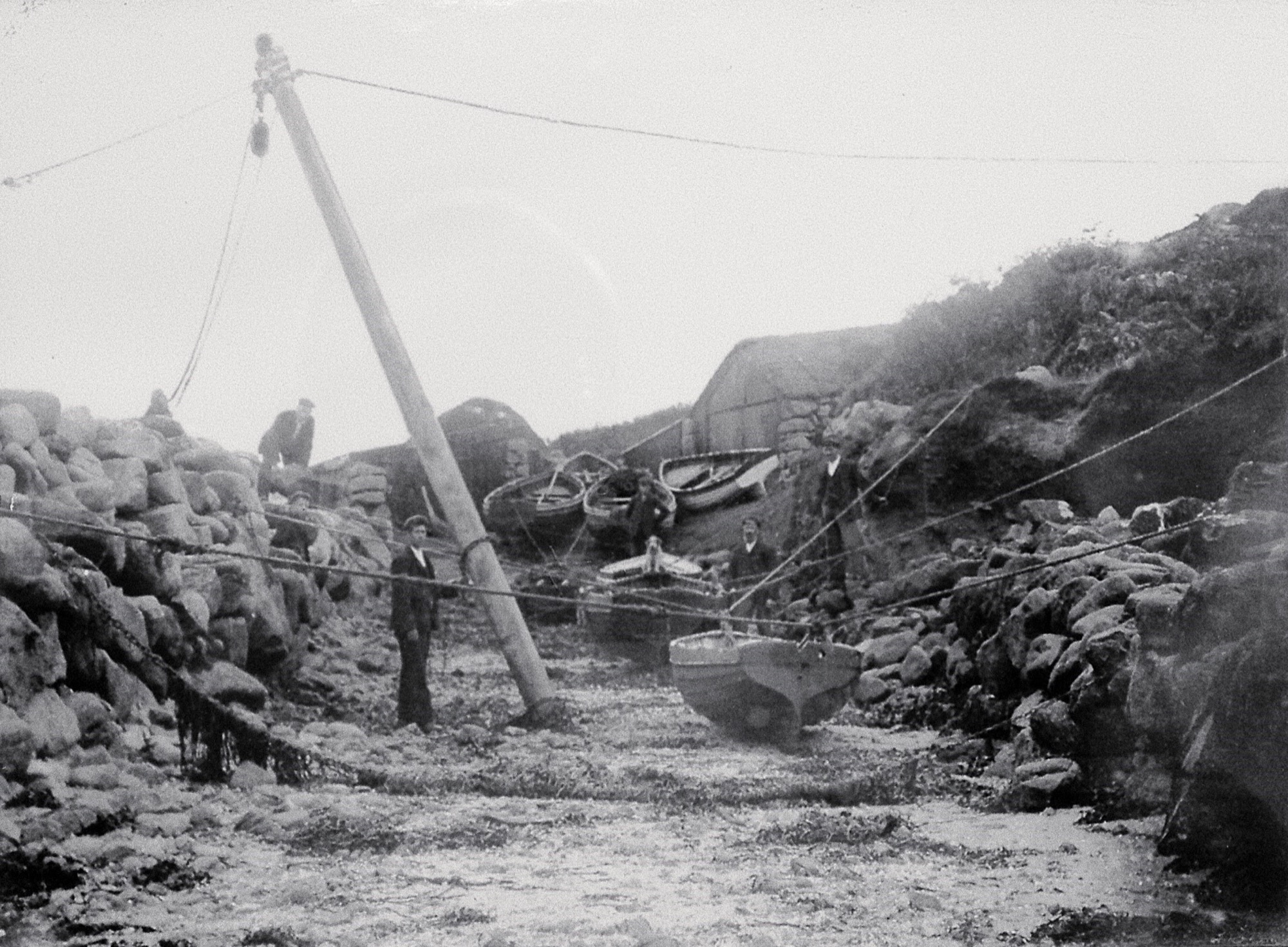 A historical black-and-white photograph of a granite-lined slipway with several people standing among moored boats. A wooden pole with ropes and pulleys is set up for lifting stones, with granite walls and gig sheds in the background.