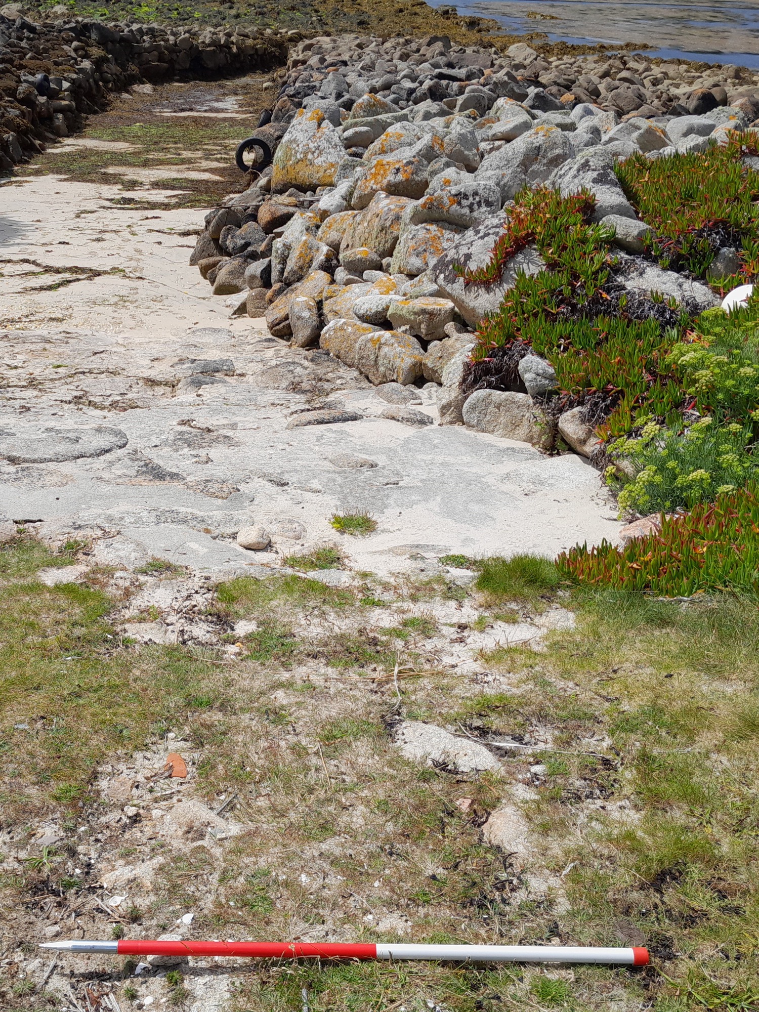 A red-and-white survey pole lies on the ground above a stone-paved slipway leading to the water. The slipway is bordered by a dry-stone revetment covered in lichen and succulent plants, with a black tyre visible further along the shore.