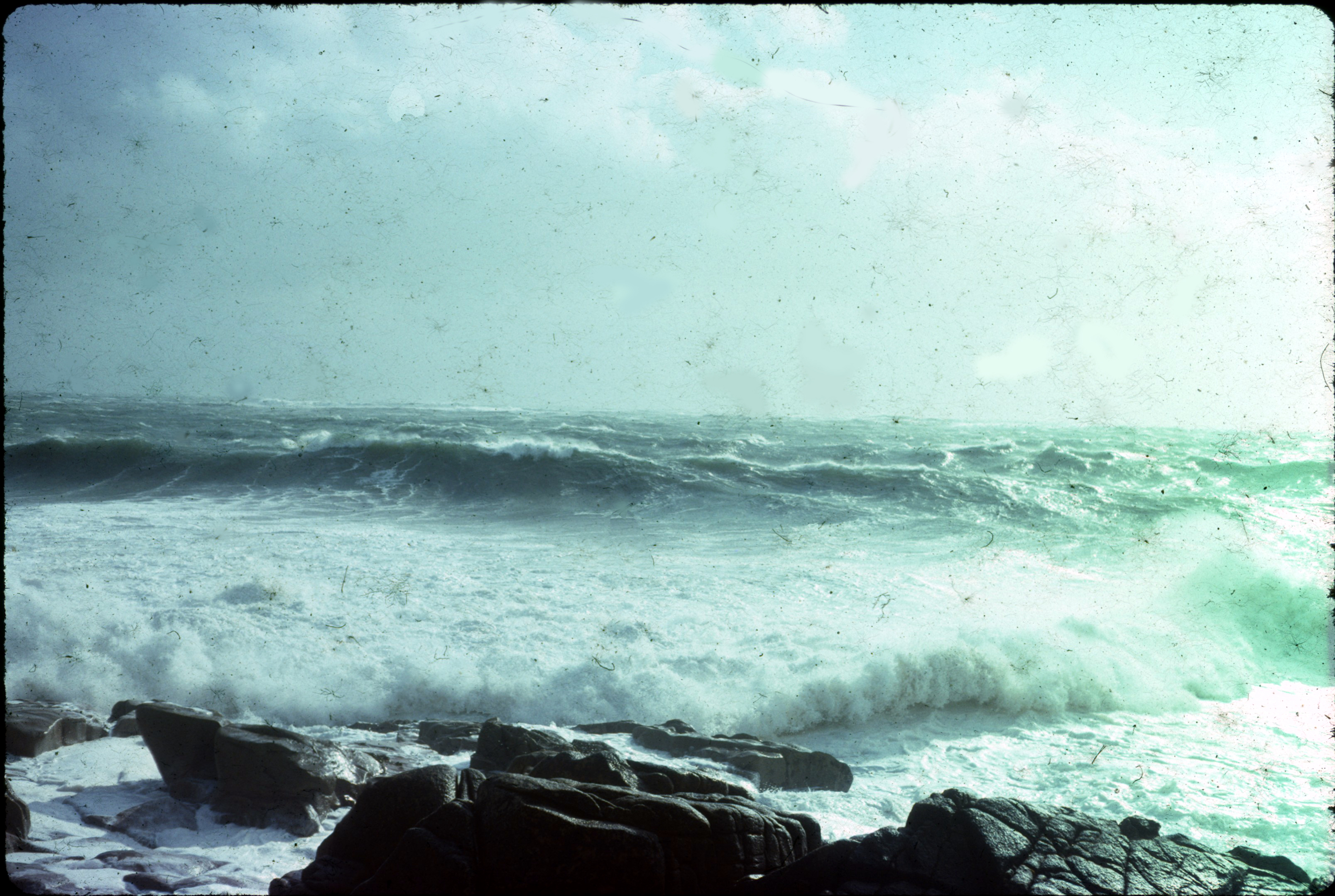 The view facing out to sea is full of large breakers with white streaked sides and curling white tops. Signs of spray in the scene and wear on the print contribute to a sense of the conditions often prevailing on the site and the passage of time since its designation.