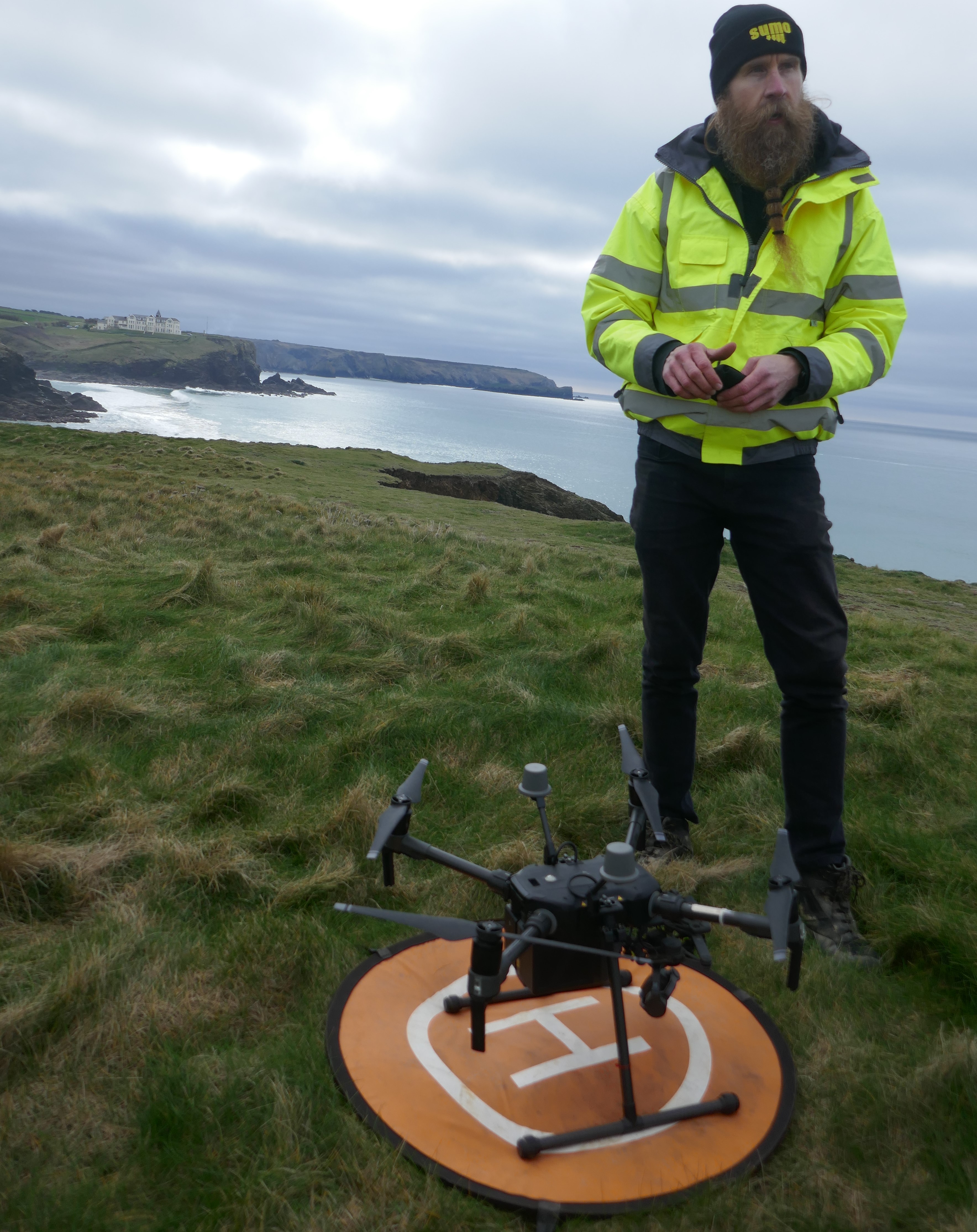A spidery black drone, almost knee height, stands on a white H on a miniature orange landing pad. Behind is the rough grassy headland and grey sea and sky, with a surveyor holding the drone control standing out in a high visibility luminous yellow coat.