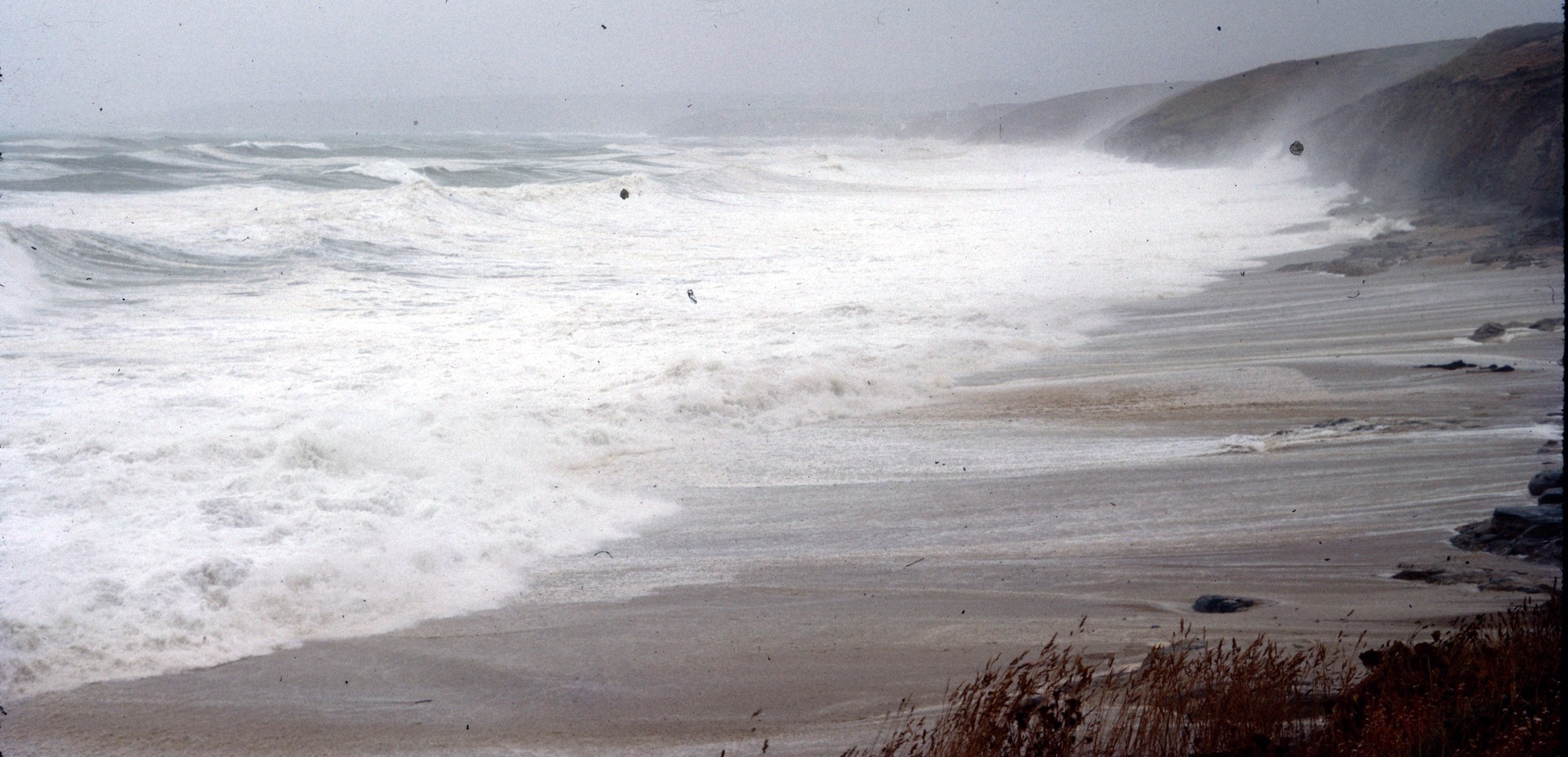 The view is almost filled by a broad zone of white surf. The shelving sands above are lined by white foam. Cliffs run away behind into the grey sky. Bits of sand are flying in the air.