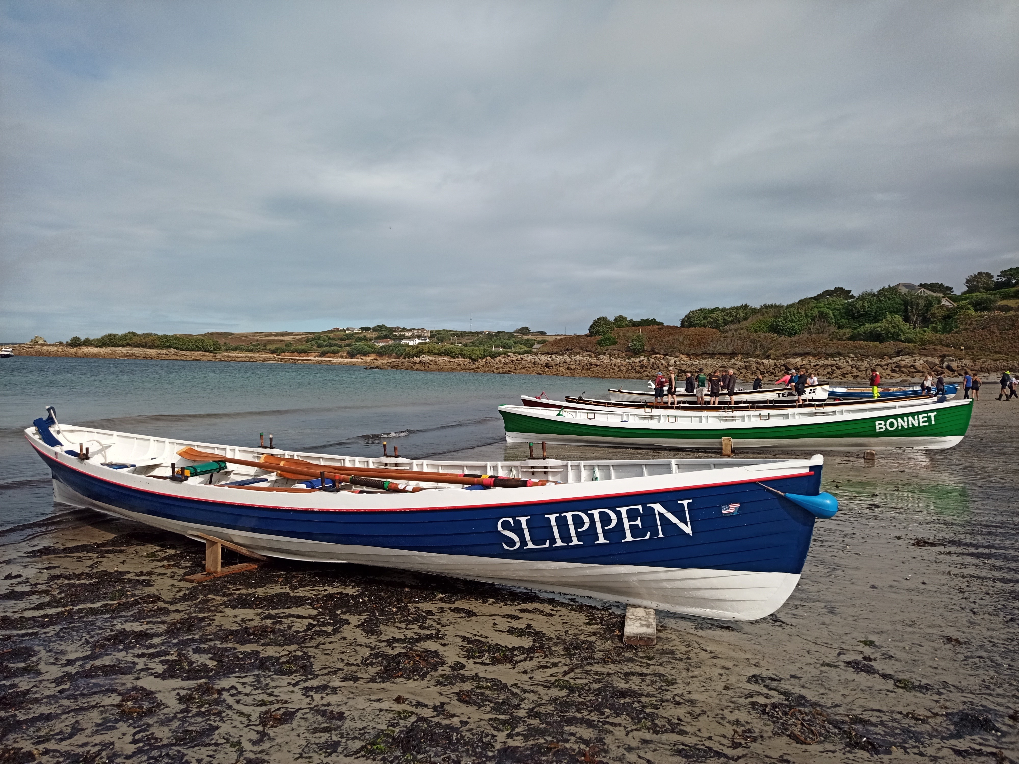 These two early gigs, Slippen painted in blue, and Bonnet in green, are drawn up on the wide sandy beach beside a calm sea at Porthmellon, St Mary’s. Beyond are Padstow gig Teaser and other boats and rowers.