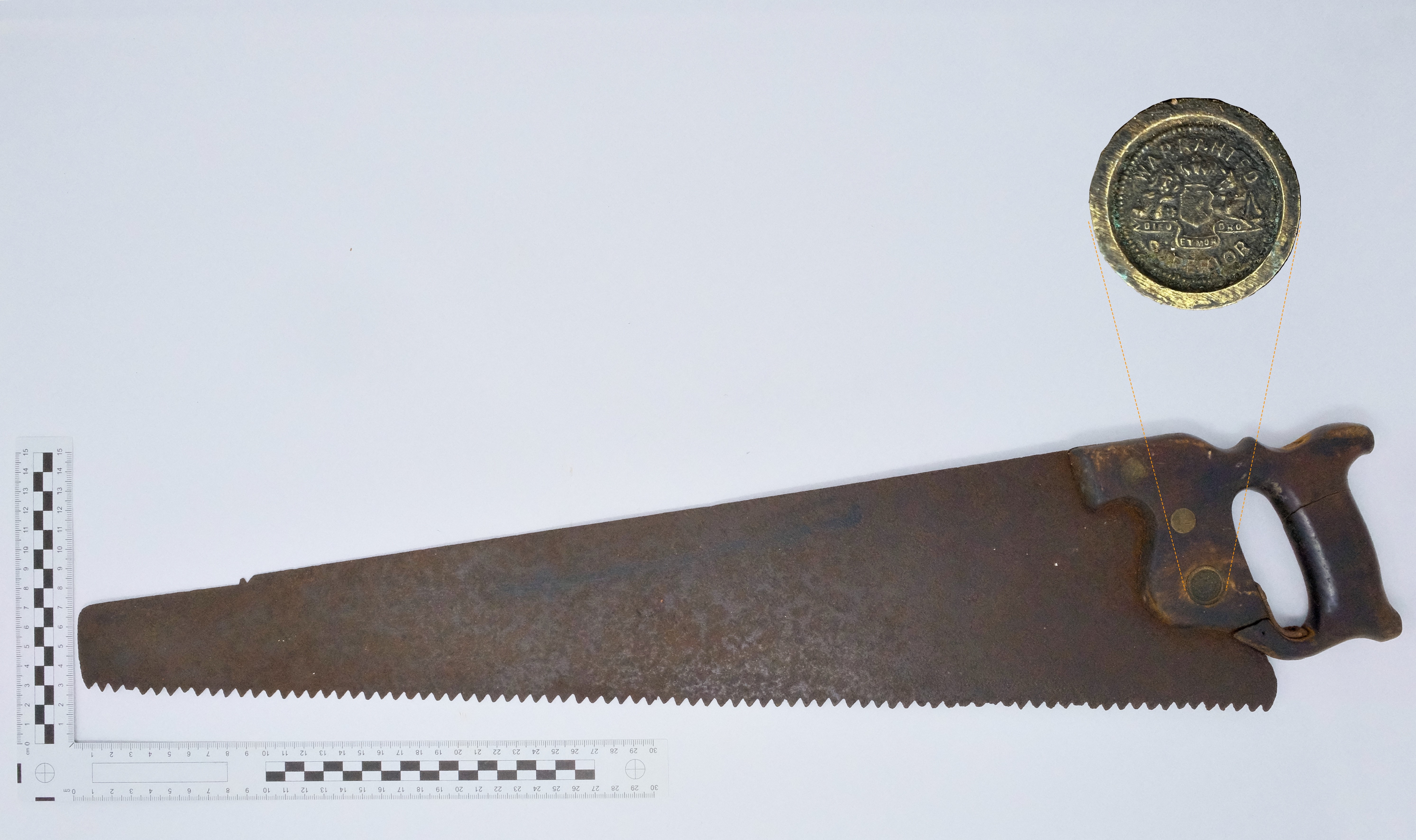 Photo of a rusty wood saw with a wooden handle, blade pointing towards the left, on a white background with a black and white scale bar on the lower left suggesting that the saw is 0.7m long. The photo contains an inset image to enlarge the circular maker’s mark ‘Warrented Superior’. 