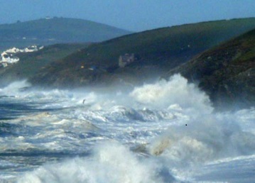 The photo is blurry with spray in the air. It shows the sea filled with breakers, crashing on a sequence of dark headlands. In the distance is a group of white houses on the outskirts of Porthleven.