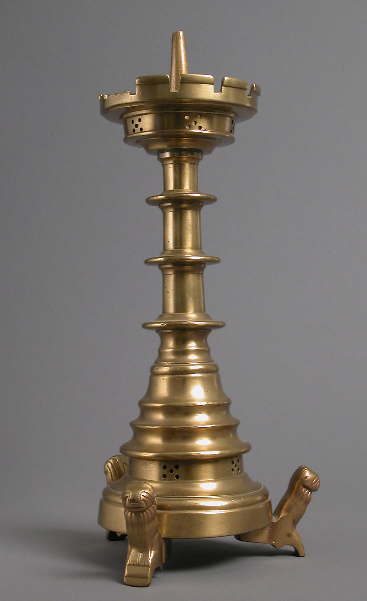 The candlestick is golden, polished brass. It is highly ornamental, with a splayed base and a broad top with crenelations like a turret around the candle spike. The three small stylised lions holding up its round base have engraved detail representing manes and sleepy faces.