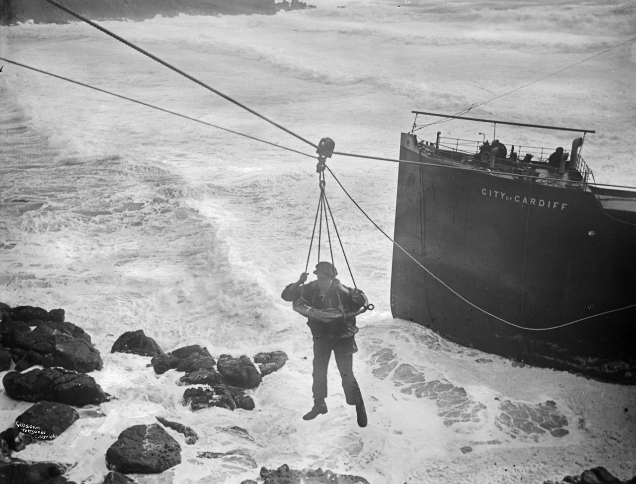 The scene is an expanse of stormy white surf foam, with the dark square bulk of the stranded steamer's bow to the right. In the centre a seaman wearing working clothes, in the breeches buoy, hangs over the sea from ropes rising from the ship to the cliff you can sense behind you from the steepness of the ropes' slant.