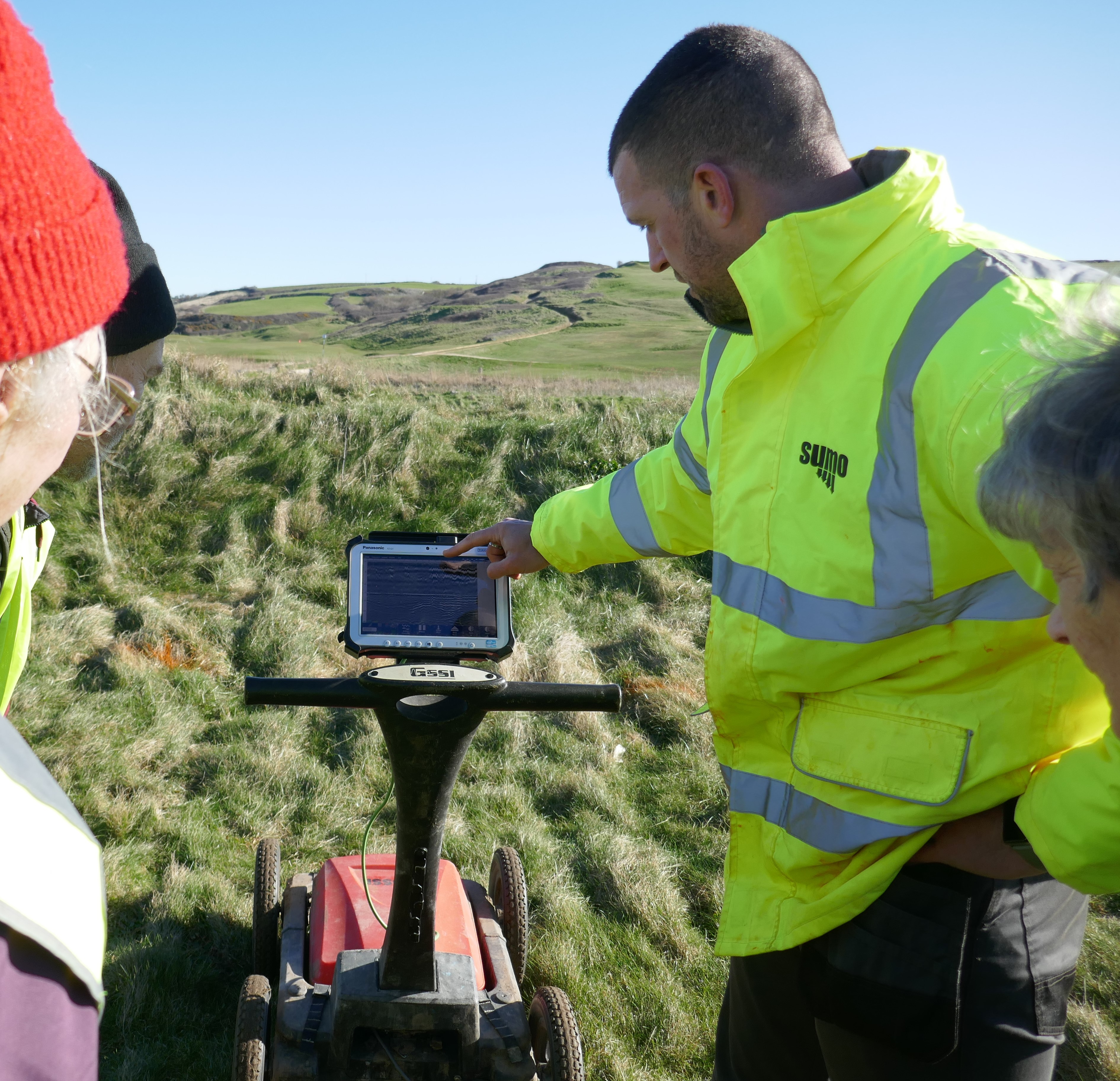 Volunteers, framing the view in the photo, are gathered round the GPR unit on the survey site. The unit has a display screen the size of a tablet and a powered base with four wheels. Beyond the site, the towans now forming a golf course rise to the horizon.