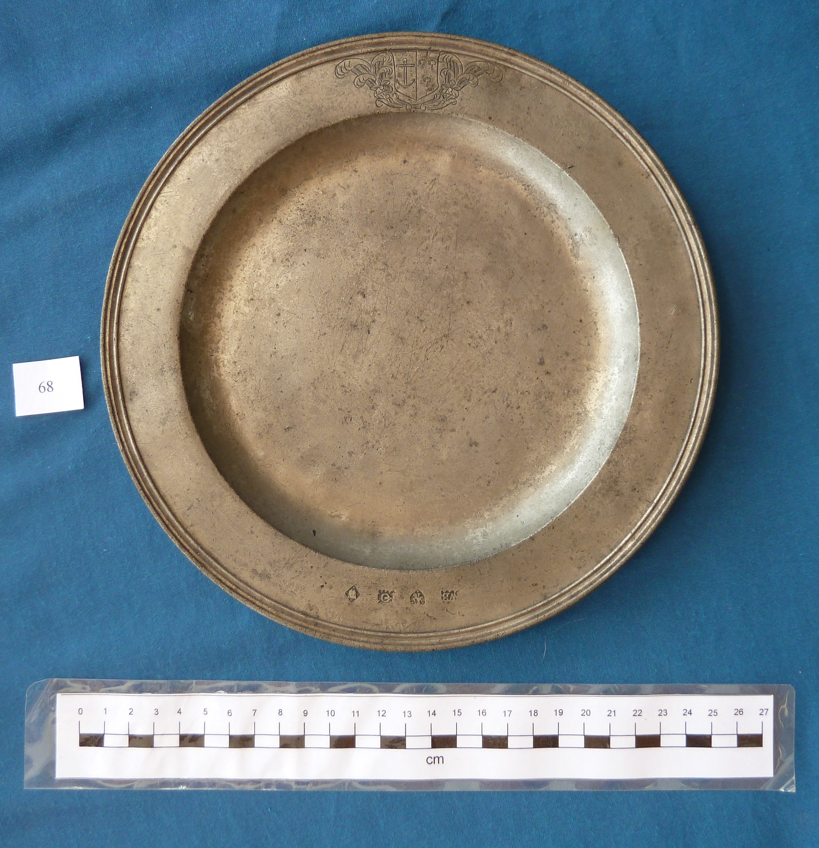 This upper surface of the plate has an enhanced, ridged edge, and a broad rim. The rim bears a coat of arms featuring an anchor engraved to one side, and opposite this across the plate, a row of four different small stamped symbols. The knife marks in the centre are quite fine but fairly numerous. The scale shows the plate is about 24cm in diameter.