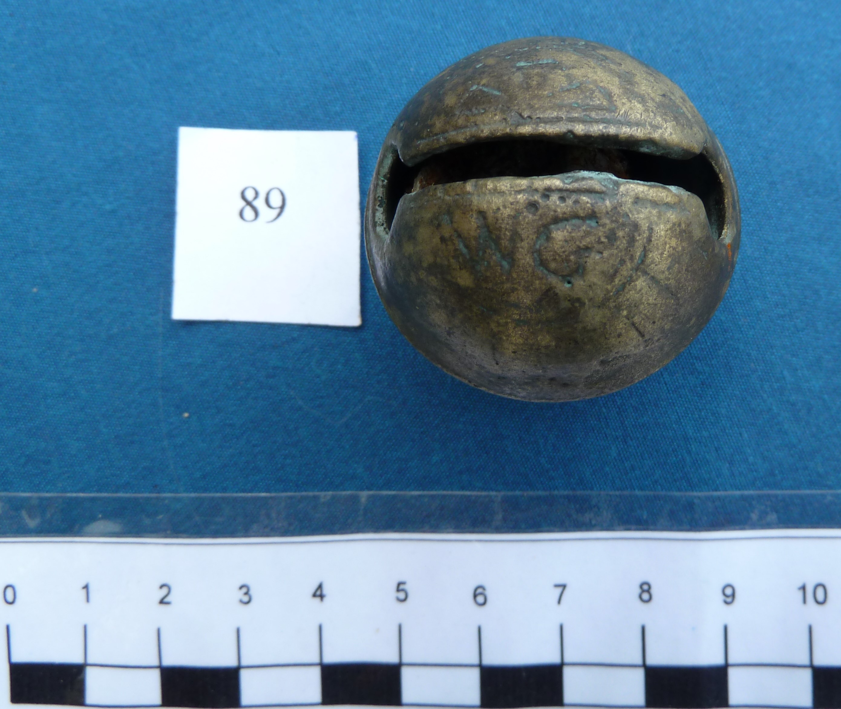 A round bell with a slit opening and some engraved lettering and patterning worn by the sea. A scale shows it is less than 5cm across.